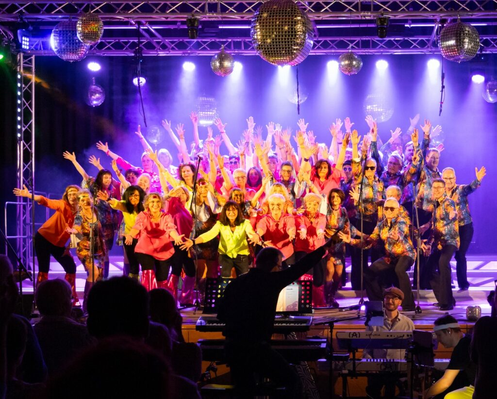 Der Chor "More Than Voices" in Disco-Outfit auf der Bühne der Ohrenberghalle unter der Leitung von Özer Dogan.